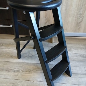 Peut inclure: Un tabouret en bois noir avec une assise ronde et trois marches. Le tabouret est pliable pour le rangement. L'assise est percée d'une poignée. Le bois est de couleur sombre.
