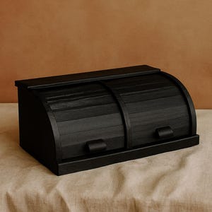 Op de afbeelding: Een zwarte houten broodtrommel met een roldeksel. De broodtrommel heeft een rechthoekige basis en een gebogen bovenkant, met een handvat aan de voorkant. De broodtrommel staat op een beige ondergrond.