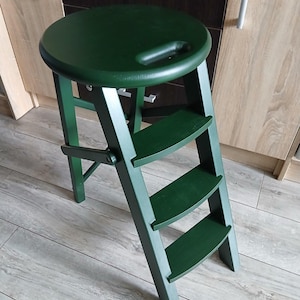Op de afbeelding: Een donkergroene houten opstap met een ronde zitting en drie treden. De kruk heeft een handvat in de zitting en is ontworpen om op te vouwen voor opslag. De treden zijn gelijkmatig verdeeld en de poten zijn schuin.