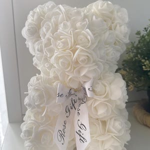 May include: A white teddy bear sculpture made from small foam roses. The bear has a white ribbon tied around its neck with the words "Rose Gift" printed on it. A decorative item.