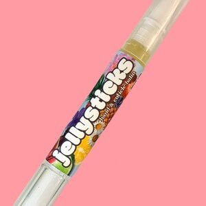 May include: A clear hand and cuticle balm pen with a white applicator tip and a clear cap. The pen's label features the text "Jellysticks hand & cuticle balm" with a colorful floral and fruit design against a pink background.