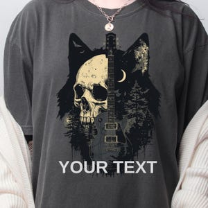 May include: Dark gray t-shirt featuring a skull, guitar, and wolf design. The graphic includes a skull with a guitar running down the center, set against a forest and crescent moon. The text "YOUR TEXT" is printed at the bottom.