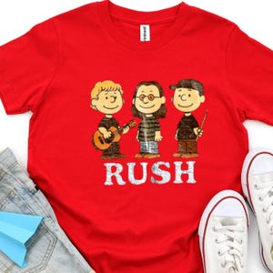 Peut inclure: T-shirt rouge avec une illustration de dessin anim&eacute; de trois musiciens et le mot "RUSH" en blanc. Le t-shirt est port&eacute; avec un jean bleu et des baskets blanches. Image d'un groupe, probablement pour un fan de musique.