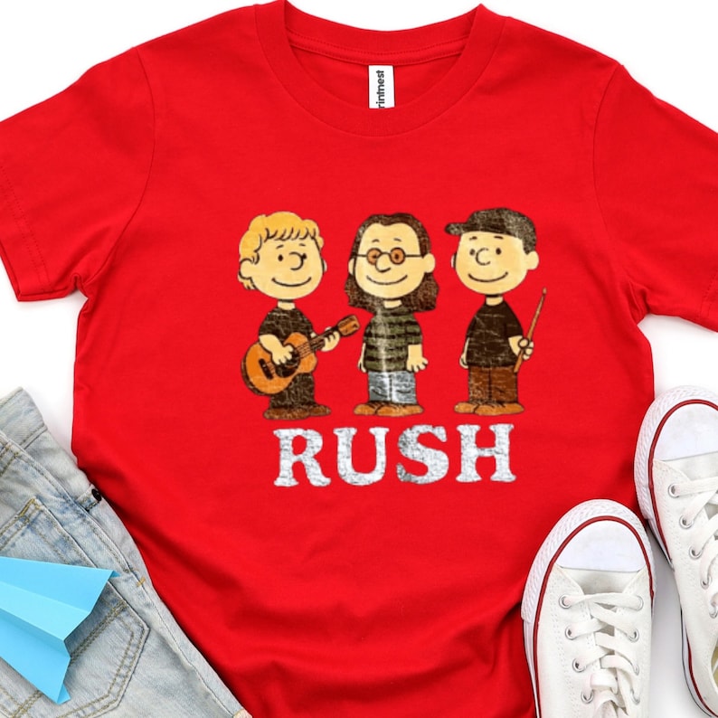 Peut inclure: T-shirt rouge avec une illustration de dessin anim&eacute; de trois musiciens et le mot "RUSH" en blanc. Le t-shirt est port&eacute; avec un jean bleu et des baskets blanches. Image d'un groupe, probablement pour un fan de musique.