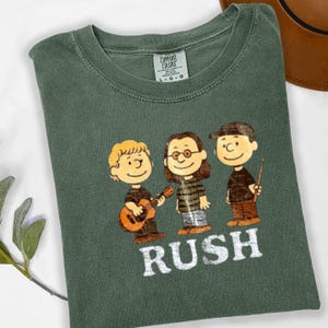 May include: Green t-shirt featuring a cartoon illustration of three characters resembling the Peanuts gang, each with a musical instrument or conductor's baton. The word "RUSH" is printed in white block letters below the characters.