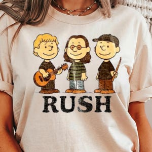 May include: Cream-colored t-shirt featuring a cartoon illustration of three characters, each holding a musical instrument. The word "RUSH" is printed in bold, black letters below the characters. The characters are wearing black shirts and brown shoes.