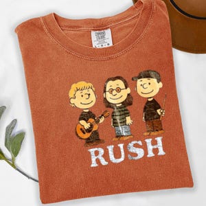 Peut inclure: T-shirt rouille avec une illustration de groupe de dessin anim&eacute; et le mot "RUSH" en lettres blanches. Les membres du groupe sont repr&eacute;sent&eacute;s dans un style vintage, l'un d'eux jouant de la guitare. Le t-shirt a un col rond et une &eacute;tiquette Comfort Colors.