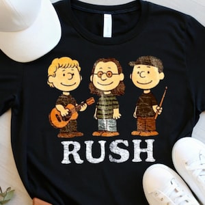 May include: Black t-shirt featuring a cartoon band with a guitar and drumsticks. The band members are styled in a cartoonish manner. The word "RUSH" is printed in white block letters below the band. A white baseball cap and white sneakers are visible.