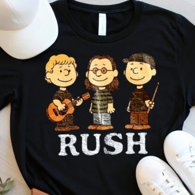 May include: Black t-shirt featuring a cartoon band with a guitar and drumsticks. The band members are styled in a cartoonish manner. The word "RUSH" is printed in white block letters below the band. A white baseball cap and white sneakers are visible.