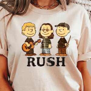 Peut inclure: T-shirt cr&egrave;me avec une illustration de dessin anim&eacute; de trois personnages jouant des instruments de musique. Le mot "RUSH" est imprim&eacute; dans une police vieillie sous les personnages. Les personnages portent des chemises noires et des pantalons marron.