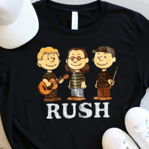Peut inclure: T-shirt noir avec une illustration de dessin anim&eacute; de trois personnages jouant d'instruments de musique. Le mot "RUSH" est imprim&eacute; en lettres blanches en dessous des personnages. Une casquette de baseball blanche et des baskets blanches sont visibles.
