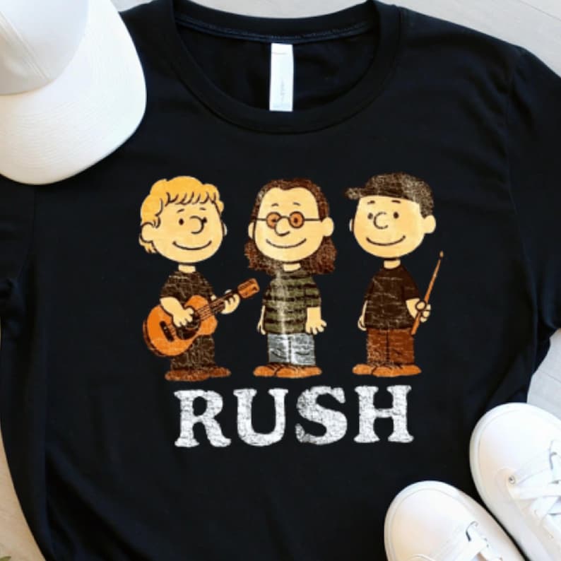 Peut inclure: T-shirt noir avec une illustration de dessin anim&eacute; de trois personnages jouant d'instruments de musique. Le mot "RUSH" est imprim&eacute; en lettres blanches en dessous des personnages. Une casquette de baseball blanche et des baskets blanches sont visibles.