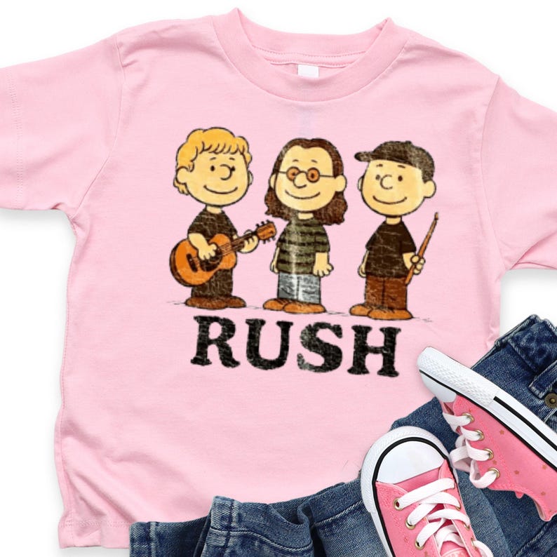 Peut inclure: T-shirt rose clair avec une illustration de dessin anim&eacute; de trois personnages jouant de la musique et le mot "RUSH" en dessous. Le t-shirt est associ&eacute; &agrave; un jean bleu et des baskets roses.