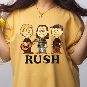 Peut inclure: T-shirt jaune moutarde avec une illustration de dessin anim&eacute; de trois personnages, dont un jouant de la guitare, et le mot "RUSH" en noir. Les personnages portent des chemises noires et des pantalons marron.