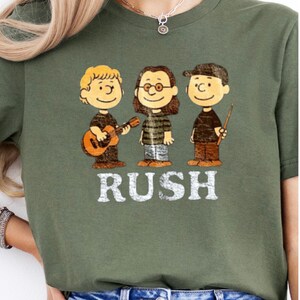 Peut inclure: T-shirt vert olive avec un motif de trois personnages de dessins anim&eacute;s rappelant le groupe Rush. Le mot "RUSH" est imprim&eacute; en blanc sous les personnages. Le personnage de gauche tient une guitare. V&ecirc;tement de style d&eacute;contract&eacute;.