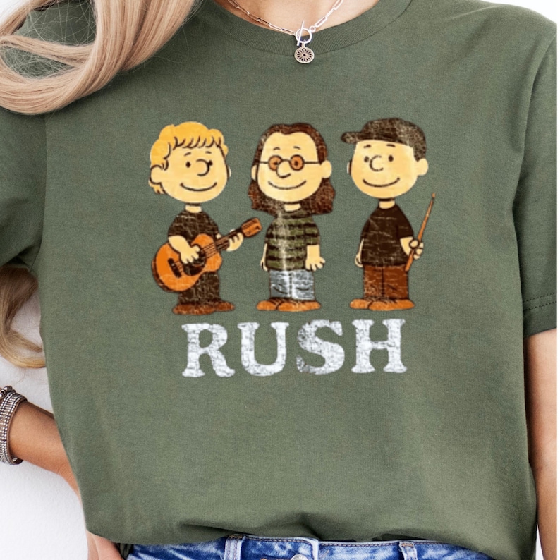 Peut inclure: T-shirt vert olive avec un motif de trois personnages de dessins anim&eacute;s rappelant le groupe Rush. Le mot "RUSH" est imprim&eacute; en blanc sous les personnages. Le personnage de gauche tient une guitare. V&ecirc;tement de style d&eacute;contract&eacute;.