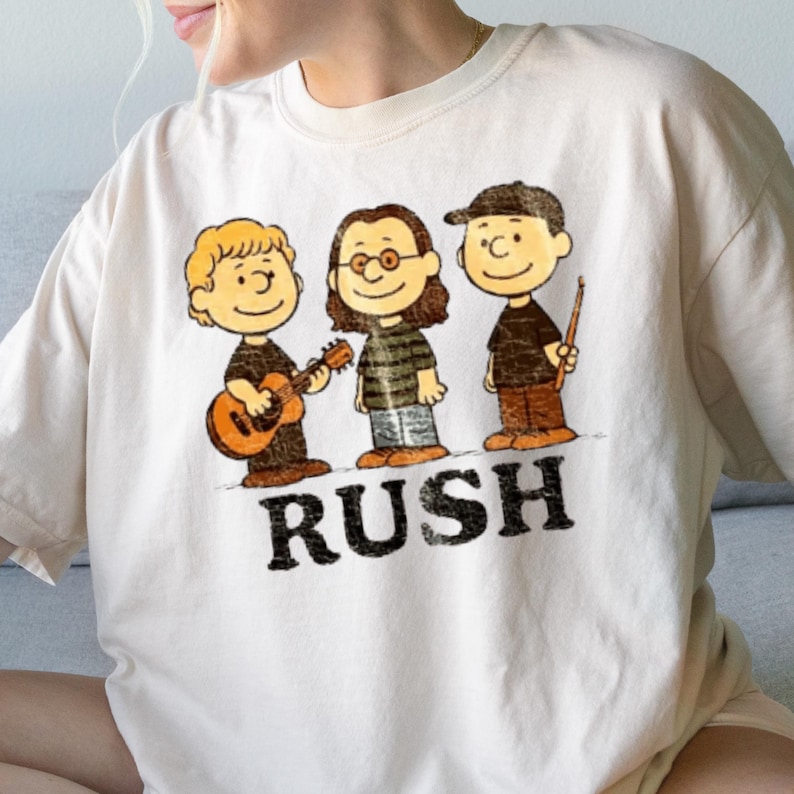 Peut inclure: T-shirt cr&egrave;me avec une illustration de dessin anim&eacute; de trois personnages jouant des instruments de musique. Le mot "RUSH" est imprim&eacute; en lettres noires en dessous des personnages. Les personnages portent des chemises noires et marron.