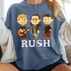 May include: A blue crew neck t-shirt featuring a cartoon illustration of three characters, each representing a member of the band Rush. The word "RUSH" is printed in white block letters below the characters.