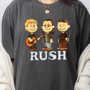Peut inclure: T-shirt gris fonc&eacute; avec une illustration de dessin anim&eacute; de trois personnages jouant des instruments de musique. Le mot "RUSH" est imprim&eacute; en lettres majuscules blanches sous les personnages. Les personnages sont marron, noir et beige.