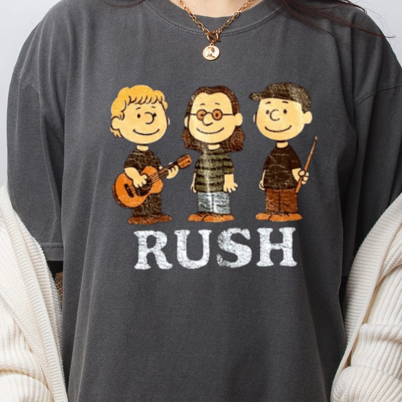 Peut inclure: T-shirt gris fonc&eacute; avec une illustration de dessin anim&eacute; de trois personnages jouant des instruments de musique. Le mot "RUSH" est imprim&eacute; en lettres majuscules blanches sous les personnages. Les personnages sont marron, noir et beige.