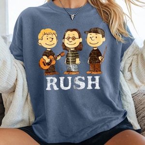 Peut inclure: T-shirt bleu avec une illustration de dessin anim&eacute; de trois personnages jouant des instruments de musique. Le mot "RUSH" est imprim&eacute; en lettres blanches en dessous. Les personnages portent des v&ecirc;tements noirs et marron.