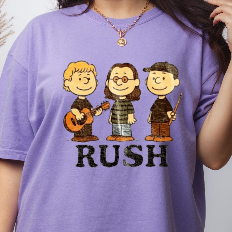 Peut inclure: Un t-shirt lilas avec une illustration de dessin anim&eacute; de trois musiciens. Les musiciens portent des chemises noires et des pantalons marron. Le mot "RUSH" est imprim&eacute; en lettres noires en dessous. L'un tient une guitare, un autre porte des lunettes et le troisi&egrave;me porte une casquette.