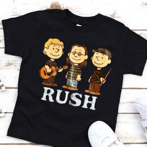 Peut inclure: T-shirt noir avec une illustration de dessin anim&eacute; de trois personnages jouant des instruments de musique. Le mot "RUSH" est imprim&eacute; en lettres blanches en dessous des personnages. Les personnages sont dans le style de la bande dessin&eacute;e Peanuts.