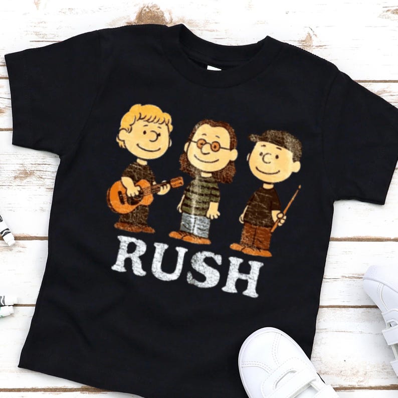 Peut inclure: T-shirt noir avec une illustration de dessin anim&eacute; de trois personnages jouant des instruments de musique. Le mot "RUSH" est imprim&eacute; en lettres blanches en dessous des personnages. Les personnages sont dans le style de la bande dessin&eacute;e Peanuts.