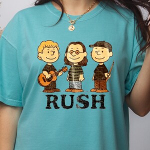 Peut inclure: T-shirt turquoise avec une illustration de dessin anim&eacute; de trois musiciens. Le mot "RUSH" est imprim&eacute; en noir en dessous. Les personnages portent des chemises noires et des pantalons marron. V&ecirc;tement de style d&eacute;contract&eacute;.