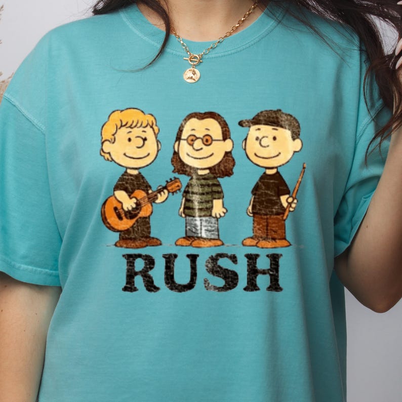 Peut inclure: T-shirt turquoise avec une illustration de dessin anim&eacute; de trois musiciens. Le mot "RUSH" est imprim&eacute; en noir en dessous. Les personnages portent des chemises noires et des pantalons marron. V&ecirc;tement de style d&eacute;contract&eacute;.