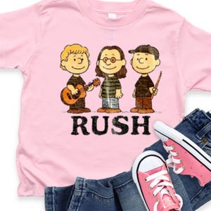 May include: Pink t-shirt featuring a cartoon band with the word "RUSH" in black block letters. The band members are styled after Peanuts characters. The shirt is paired with blue jeans and pink sneakers.