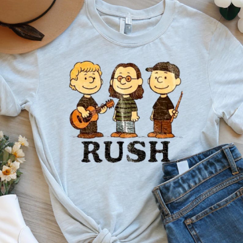 May include: Light blue t-shirt featuring a cartoon illustration of three characters playing musical instruments. The word "RUSH" is printed in black block letters below the characters. The shirt is paired with blue jeans and a brown hat.