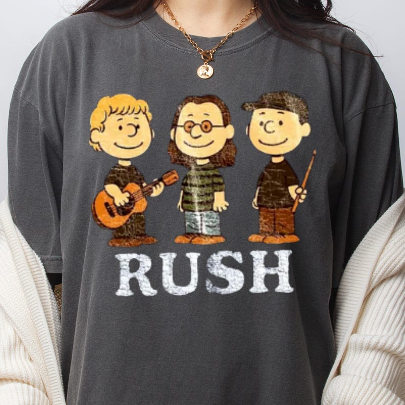 May include: Dark gray t-shirt featuring a cartoon illustration of three characters playing musical instruments. The word "RUSH" is printed in white block letters below the characters. The characters are in the style of the Peanuts comic strip.