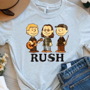 Peut inclure: T-shirt bleu clair avec une illustration de dessin anim&eacute; de trois musiciens et le mot "RUSH" en noir. Les personnages jouent de la guitare, portent des lunettes et tiennent des baguettes. Un chapeau marron et un jean bleu sont partiellement visibles.