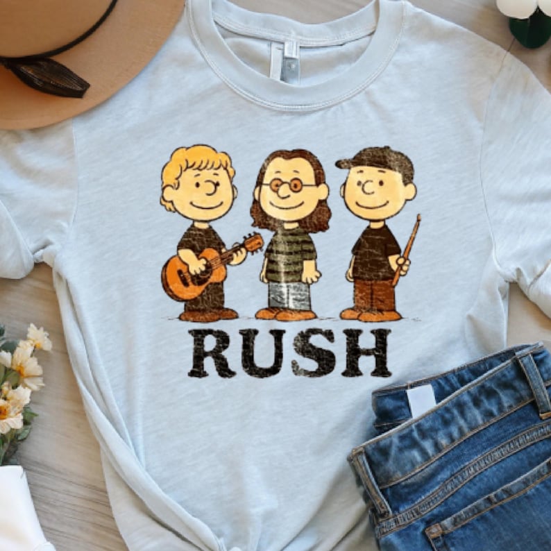 Peut inclure: T-shirt bleu clair avec une illustration de dessin anim&eacute; de trois musiciens et le mot "RUSH" en noir. Les personnages jouent de la guitare, portent des lunettes et tiennent des baguettes. Un chapeau marron et un jean bleu sont partiellement visibles.