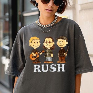 Peut inclure: T-shirt gris fonc&eacute; avec une illustration de dessin anim&eacute; de trois musiciens et le mot "RUSH" en blanc. Les musiciens de dessins anim&eacute;s comprennent un jouant de la guitare, un autre portant des lunettes et le troisi&egrave;me tenant des baguettes.