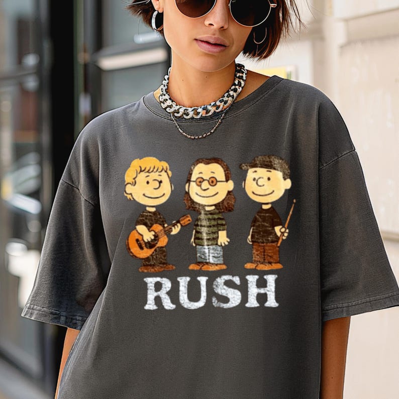 Peut inclure: T-shirt gris fonc&eacute; avec une illustration de dessin anim&eacute; de trois musiciens et le mot "RUSH" en blanc. Les musiciens de dessins anim&eacute;s comprennent un jouant de la guitare, un autre portant des lunettes et le troisi&egrave;me tenant des baguettes.