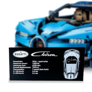 Op de afbeelding: Een zwart rechthoekig displayplaatje voor een Bugatti Chiron modelauto. Het plaatje bevat het Bugatti-logo, het Chiron-schrift en technische specificaties waaronder W16 quad-turbo, 1500 pk, 6700 tpm, 7993 cc, 7-traps dubbele koppeling en acceleratietijden.