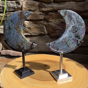 May include: Two crescent moon-shaped polished stone carvings featuring mottled blue, grey, and magenta mineral inclusions. Each stone is mounted on a metal stand, one with a black finish and the other with a silver finish, displaying the natural patterns.