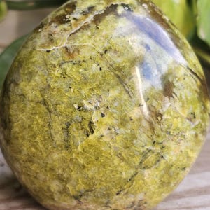 May include: A smooth, rounded stone with a mottled green and yellow surface. The stone has dark veins and a polished appearance, suggesting it is a decorative item or a natural specimen. The stone is approximately 4 inches in diameter.