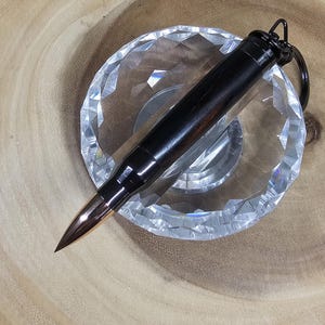 May include: A black bullet-shaped keychain with a bronze-colored tip rests on a clear, faceted glass dish. The keychain is made of dark wood and metal. The dish sits on a light brown, wooden surface.