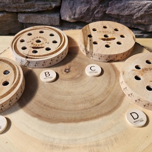 May include: Four wooden game pieces with multiple holes and the text "MERRILLY MARKET" are displayed on a wooden surface. Small wooden discs labeled A, B, C, and D are also present. The pieces have a natural wood grain.