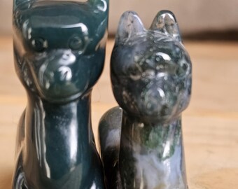 Puede incluir: Dos figuras de gato talladas en piedra. El gato de la izquierda es de color verde oscuro, mientras que el gato de la derecha es una mezcla de gris, blanco y verde. Ambos gatos están de pie con las orejas erguidas.