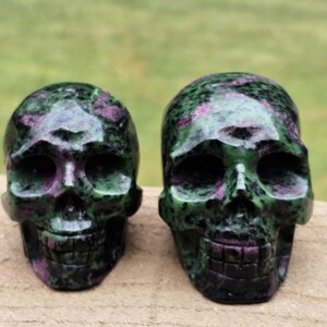 May include: Two carved stone skulls. The skulls are green with pink and black veining. The skulls are displayed on a light brown surface with a blurred green background.