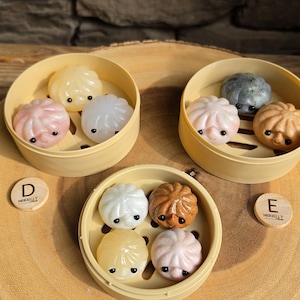 May include: A collection of colorful, dumpling-shaped objects in small bamboo steamer baskets. The dumplings are in shades of pink, white, yellow, brown, and gray. Each dumpling has small black eyes. The scene is set on a wooden surface.