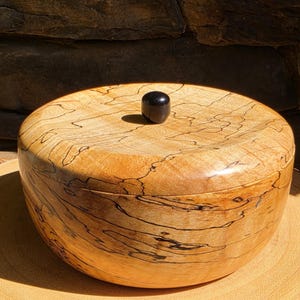 May include: A circular wooden box with a lid and a small, dark knob. The box is crafted from light-coloured wood with dark, swirling grain patterns. The box rests on a wooden surface.