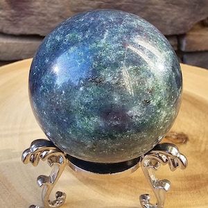 May include: A polished, spherical gemstone in shades of blue and green, resting on a decorative silver stand. The sphere's surface reflects light, showcasing its natural patterns. The stand has an ornate, stylized design.