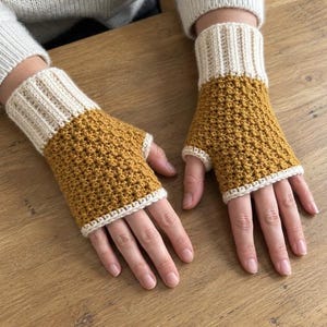 May include: A pair of fingerless gloves featuring a mustard yellow textured crochet body and a ribbed cream-colored cuff. The gloves have a simple opening for the thumb and a finished cream-colored edge around the fingers and thumb hole.