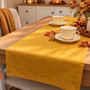 Peut inclure: Un chemin de table rectangulaire jaune moutarde en tissu texturé, posé à plat sur une table à manger en bois. Le chemin de table présente un bord ourlé simple et un aspect tissé, offrant une base de couleur unie pour la décoration de table.