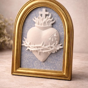 May include: A white heart sculpture with a crown of thorns and a cross, set against a blue patterned background. The heart is framed in a gold-coloured, arched frame. The artwork is a religious symbol.